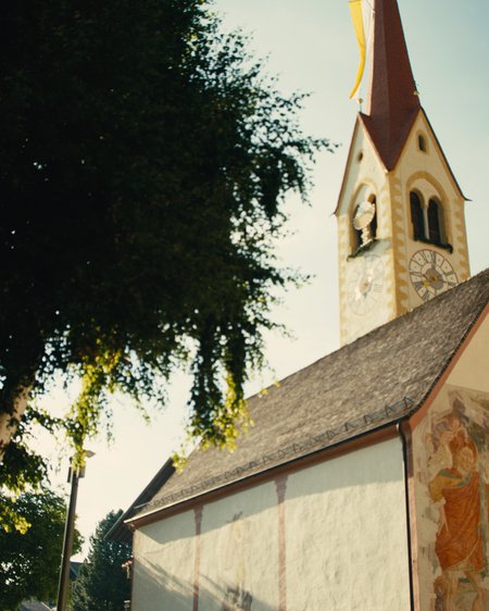 Unser Hotel in Olang zwischen Himmel und Erde Kirche mit Uhr und bemalter Fassade neben großen Bäumen bei Sonnenlicht