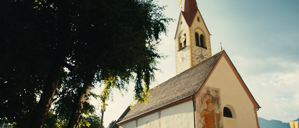 Unser Hotel in Olang zwischen Himmel und Erde Kirche mit Uhr und bemalter Fassade neben großen Bäumen bei Sonnenlicht