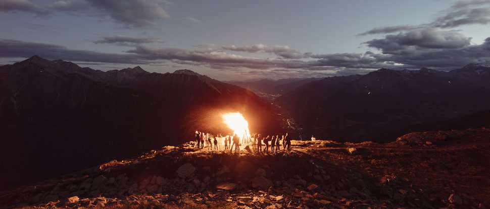 Unser Hotel in Olang zwischen Himmel und Erde Menschen um ein großes Lagerfeuer auf einem Berggipfel bei Sonnenuntergang