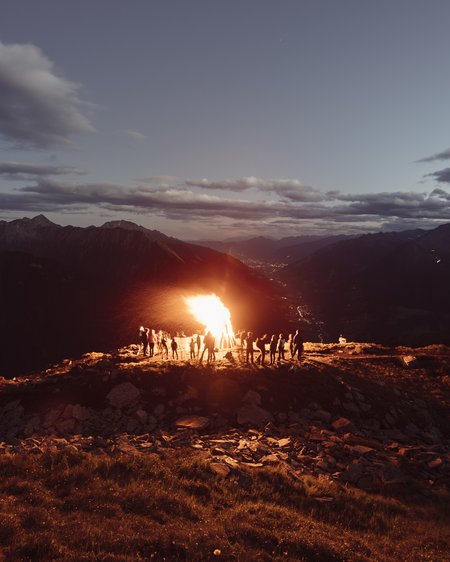 Unser Hotel in Olang zwischen Himmel und Erde Menschen um ein großes Lagerfeuer auf einem Berggipfel bei Sonnenuntergang