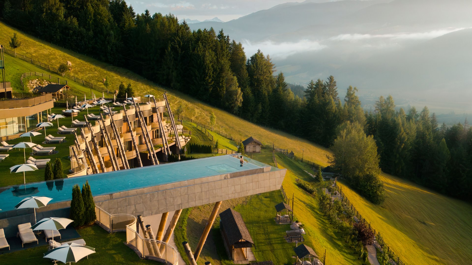 Unser Hotel in Olang zwischen Himmel und Erde Infinity-Pool auf Holzstützen an Berghang mit Wald und Bergen im Hintergrund