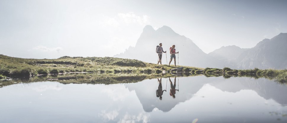 Unser Hotel in Olang zwischen Himmel und Erde Zwei Wanderer mit Rucksäcken neben einem Bergsee mit Bergreflexion