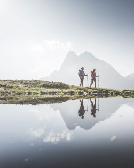Unser Hotel in Olang zwischen Himmel und Erde Zwei Wanderer mit Rucksäcken neben einem Bergsee mit Bergreflexion
